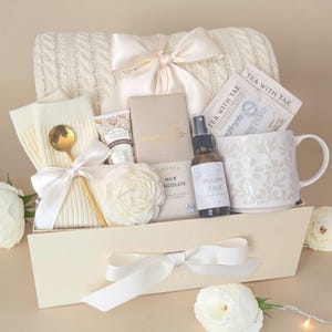 May include: A gift set in a cream-colored box featuring a cream cable-knit blanket with a satin bow, a floral-patterned mug, a Tea With Tae tea bag, a milk chocolate bar, a small spray bottle labeled Pillow Talk, a gold spoon, and a white flower.
