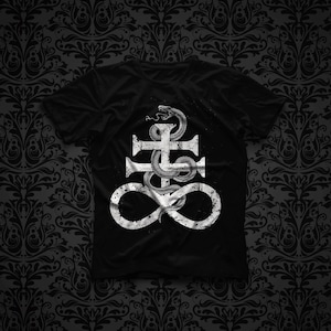 Può includere: Una t-shirt nera a maniche corte con una stampa grafica bianca di un serpente avvolto attorno al simbolo della croce di Leviatano. Il design presenta una texture usurata e invecchiata, centrata sul petto del capo con scollo rotondo.