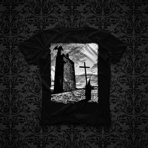 Puede incluir: Una camiseta negra de manga corta con un estampado gráfico blanco de alto contraste en el pecho. El diseño muestra una torre de piedra alta con una figura alada en la parte superior, junto a una gran cruz y una figura encapuchada en un paisaje.