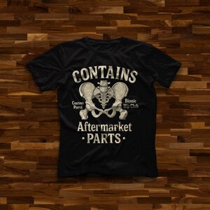 May include: A black short-sleeved t-shirt featuring a beige anatomical illustration of a human pelvis in the center. The text reads CONTAINS, Custom Parts, Bionic Hip Club, AFTERMARKET, and PARTS in a distressed, vintage-style beige font.