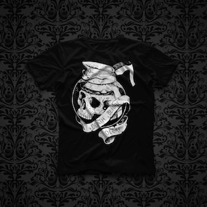May include: A black t-shirt featuring a white graphic print of a human skull inside a glass coffee carafe with a lid. A ribbon banner wraps around the carafe with the text DEATH BEFORE DECAF printed in black capital letters on the white ribbon.
