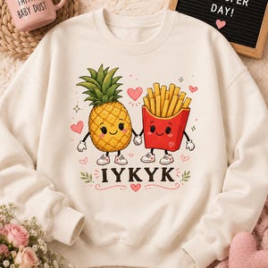 Puede incluir: Una sudadera de cuello redondo color crema con un gráfico de una piña y una caja de patatas fritas de dibujos animados tomados de la mano. El diseño incluye pequeños corazones rosas, destellos y el texto IYKYK en letras negras negritas.
