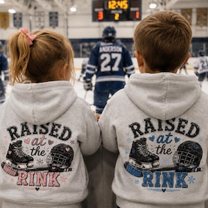 May include: Two light grey hooded sweatshirts featuring a graphic on the back with the text RAISED at the RINK. The design includes illustrations of black ice skates, a hockey helmet, and a hockey stick, accented with small hearts and snowflakes.