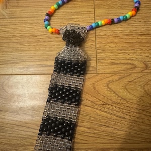 May include: A necktie crafted from plastic pony beads featuring a striped pattern of alternating black and clear beads. The neck loop is constructed from a single strand of multicolored beads in a rainbow sequence, including red, orange, yellow, green, blue, and purple.