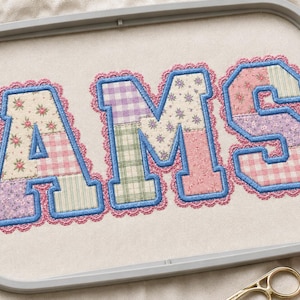 May include: The letters AMS are embroidered in a varsity font on a light fabric within a gray embroidery hoop. Each letter features a patchwork of floral, plaid, and gingham patterns in pastel colors, outlined in blue thread with a pink scalloped edge.