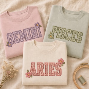 Könnte beinhalten: Drei Rundhals-Sweatshirts in Hellrosa, Salbeigrün und Creme. Jedes zeigt gestickte Buchstaben im College-Stil für die Sternzeichen GEMINI, PISCES und ARIES, gefüllt mit einem Blumenmuster und akzentuiert mit kleinen gestickten Blüten.