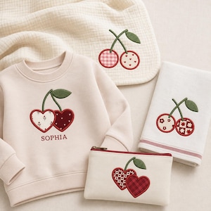 Könnte beinhalten: Ein cremefarbenes Sweatshirt mit dem Namen SOPHIA unter einem herzförmigen Kirschmotiv, dazu ein passendes cremefarbenes Reißverschlusstäschchen und ein gefaltetes cremefarbenes Tuch, beide mit gestickten herzförmigen Kirschmotiven.
