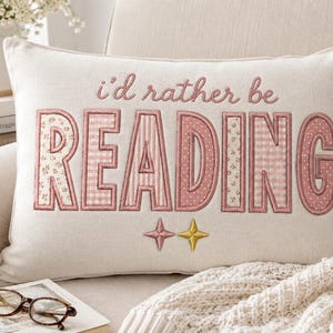 Könnte beinhalten: Ein rechteckiges, cremefarbenes Zierkissen mit dem gestickten Text i'd rather be READING in rosa Garn. Die Buchstaben sind mit verschiedenen rosa-weißen Stoffmustern wie Vichy-Karos und Blumen gefüllt, ergänzt durch zwei kleine Sternsymbole darunter.
