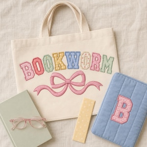 Könnte beinhalten: Eine cremefarbene Canvas-Tragetasche mit dem gestickten Text BOOKWORM in mehrfarbigen Buchstaben über einer rosa Schleife. Daneben liegen ein hellgrünes Buch, eine Brille mit rosa Rahmen, ein gelbes Lesezeichen und eine blaue, gesteppte Buchhülle.