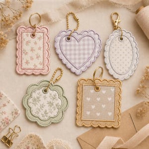 May include: Five embroidered fabric keychains with scalloped edges and metal hardware. Designs include a pink floral rectangle, a lavender gingham heart, a white polka dot oval, a sage green floral flower, and a tan square with white hearts.