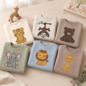 Puede incluir: Seis sudaderas de cuello redondo en varios colores pastel, cada una con un animal bordado diferente: un tigre, un mono colgado de una rama, un oso, un elefante, un león y una jirafa. Las prendas están dispuestas en un patrón de cuadrícula.