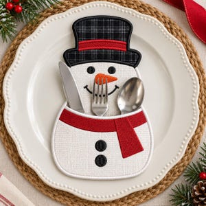 Könnte beinhalten: Ein weißer Besteckhalter aus Stoff in Form eines Schneemanns mit schwarzem Karomuster-Zylinder, orangefarbener Karottennase, rotem Schal und zwei schwarzen Knöpfen. Der Halter enthält ein silbernes Messer, eine Gabel und einen Löffel.