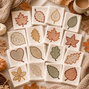 Könnte beinhalten: Eine Sammlung von sechzehn quadratischen weißen Stoffservietten, die jeweils mit einem einzigartigen gestickten Blattmotiv versehen sind. Die Blätter sind in verschiedenen herbstlich inspirierten Mustern wie Vichy, Blumen und einfarbigen Texturen in Orange-, Braun- und Grüntönen dargestellt.