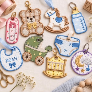 Könnte beinhalten: Eine Sammlung bestickter Schlüsselanhänger mit Babymotiven, darunter ein Teddybär, ein Schaukelpferd, ein Babyfläschchen, eine Sicherheitsnadel, ein Lätzchen mit dem Namen NOAH, ein Kinderwagen, eine Windel, ein Kinderbett und ein Mond-Wolken-Mobile.