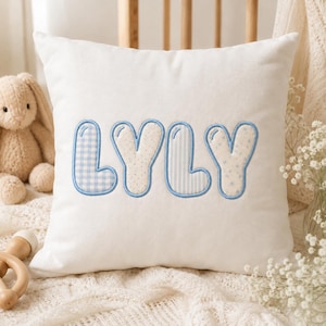 Puede incluir: Un cojín cuadrado de color blanco con el nombre LYLY bordado en letras grandes estilo burbuja. Cada letra está delineada con hilo azul y rellena con diferentes patrones en azul claro, incluyendo cuadros vichy, puntos pequeños, rayas y estrellas.