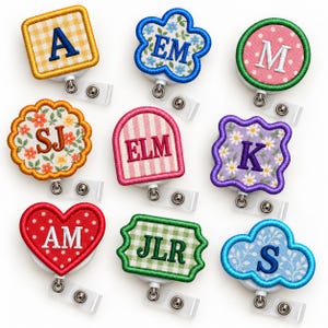 May include: A collection of nine badge reels featuring embroidered fabric patches in various shapes including squares, circles, hearts, and clouds. Each patch displays different letters or initials in colorful patterns like gingham, polka dots, and floral prints.