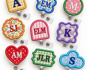 May include: A collection of nine badge reels featuring embroidered fabric patches in various shapes including squares, circles, hearts, and clouds. Each patch displays different letters or initials in colourful patterns like gingham, polka dots, and floral prints.