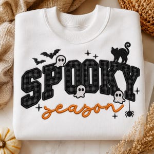 May include: A white crewneck sweatshirt featuring embroidered text that reads SPOOKY in a black and grey buffalo plaid pattern and season in orange cursive. The design includes small white ghosts, black bats, a black cat, and a spider.