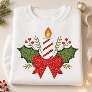 Könnte beinhalten: Ein weißes Sweatshirt mit Rundhalsausschnitt und einer Stickerei, die eine zentrale Kerze mit rot-weißen Diagonalstreifen, einer gelben Flamme, grünen Stechpalmenblättern, roten Beeren und einer großen roten Schleife mit Schneeflockenmuster zeigt.