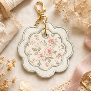 May include: A scalloped, flower-shaped fabric keyring featuring a cream-coloured base with a delicate pink rose and green leaf floral pattern. The edges are finished with sage green embroidery, and it includes a gold-toned metal lobster clasp.