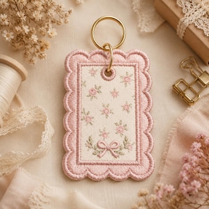 May include: A rectangular embroidered keyring with a scalloped edge in dusty pink thread. The centre features a cream-coloured fabric with a repeating pattern of small pink flowers, green leaves, and a central pink bow, attached to a gold metal ring.