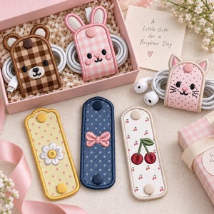 May include: A collection of embroidered cable organizers featuring animal and patterned designs. Two organizers are shaped like a bear and a rabbit, holding white cables inside a pink box. Three rectangular organizers feature daisy, bow, and cherry motifs.