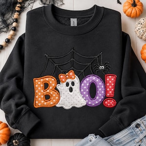 Könnte beinhalten: Ein schwarzes Sweatshirt mit Rundhalsausschnitt und einem gestickten Design, das den Text BOO! in orangefarbenen, violetten und roten gepunkteten Buchstaben zeigt. Ein weißes Gespenst mit orangefarbener Schleife und ein Spinnennetz sind darüber.