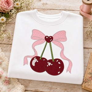 Könnte beinhalten: Ein weißes Sweatshirt mit Rundhalsausschnitt und einer zentralen Stickerei von zwei dunkelroten Kirschen mit grünen Stielen. Die Kirschen sind mit einer großen, hellrosa gepunkteten Schleife verbunden, die in der Mitte ein kleines Herz hat.