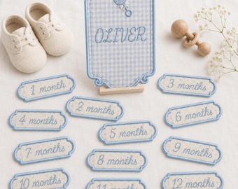 May include: A set of baby milestone markers featuring a blue and white gingham plaque with the name Oliver and a rattle icon, accompanied by twelve rectangular embroidered labels labelled 1 month through 12 months and two cream baby shoes.
