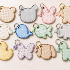 May include: A collection of twelve fabric keychains in various animal and insect shapes, including a bear, cat, rabbit, whale, fish, elephant, duck, butterfly, and dinosaur. Each keychain features patterned fabric, a metal ring, and a small charm.