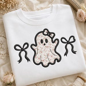 May include: A white crewneck sweatshirt featuring an embroidered ghost character with a floral pattern in shades of pink and green. The ghost wears a small bow on its head and is flanked by two black embroidered bows on either side of the design.