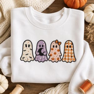 Könnte beinhalten: Ein weißes Sweatshirt mit Rundhalsausschnitt und einer Stickerei von vier Geistern in einer Reihe. Der erste Geist hat ein schwarzes Fledermausmuster, der zweite ist lila mit einem Halbmond, der dritte ist pfirsichfarben mit Kürbissen und der vierte kariert.
