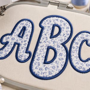 Könnte beinhalten: Ein Stickrahmen mit einem hellen Stoff, auf dem die Buchstaben A, B und C in einem blauen Blumenmuster gestickt sind. Die Buchstaben sind mit einem dicken, dunkelblauen Satinstich umrandet, was für eine erhabene, strukturierte Optik sorgt.