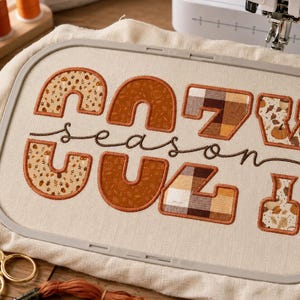 Op de afbeelding: Een borduurring met gebroken witte stof en de geborduurde tekst "cozy season". De letters zijn gevuld met diverse herfstpatronen, waaronder bruine ruiten, oranje pompoenprints en herfstbladeren in tinten van roest en crème.