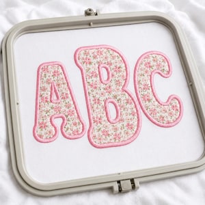 May include: A square embroidery hoop containing white fabric with the letters ABC appliqued in the center. The letters feature a floral pattern with small pink flowers and green leaves on a cream background, outlined with thick pink satin stitching.