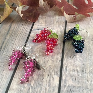 May include: Three pairs of dangle earrings featuring grape clusters. The earrings are made of small beads and have a silver tone. One pair is pink, one is red, and one is dark blue.