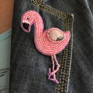 May include: Pink beaded flamingo brooch with a pink and grey stone in the center. The brooch is pinned to a denim jacket.