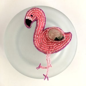 May include: Pink beaded flamingo with a pink and black stone in the center of its body. The flamingo is standing on a white plate.