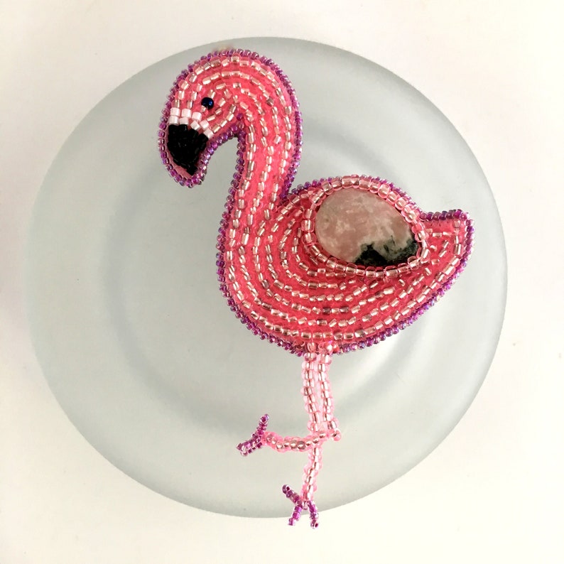 May include: Pink beaded flamingo with a pink and black stone in the center of its body. The flamingo is standing on a white plate.