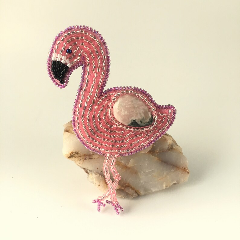 May include: A pink beaded flamingo with a pink and green stone on its back. The flamingo is standing on a white rock.