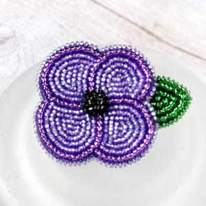 May include: A purple beaded flower with a green leaf. The flower is made of small, round beads and has a center made of black beads.