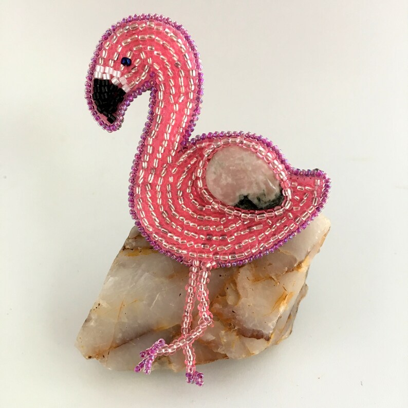 May include: Pink beaded flamingo with a pink and black stone in the center of its body. The flamingo is standing on a light brown rock.