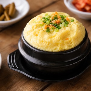 May include: A fluffy, yellow steamed egg dish served in a black ceramic bowl with a lid base. The top of the egg is garnished with finely chopped green onions, small orange carrot pieces, and a light dusting of black pepper and sesame seeds.