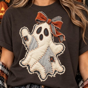 Può includere: Una t-shirt grigio antracite scuro con una grande applicazione centrale a forma di fantasma. Il fantasma è realizzato con toppe in tessuto color crema e grigio chiaro dai bordi sfrangiati, dettagli a punto croce arancione e un fiocco.