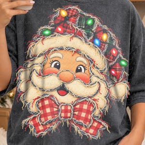 May include: A charcoal grey t-shirt featuring a graphic of Santa Claus wearing a red and green plaid hat adorned with colourful string lights. Santa has a white beard, a red and white gingham bow tie, and a distressed, frayed-edge design.