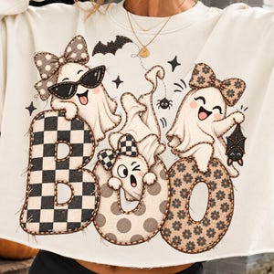 Pode incluir: Uma camisola de cor creme com um gráfico grande de três fantasmas de desenhos animados e a palavra BOO. Os fantasmas usam laços estampados e óculos de sol. As letras estão preenchidas com padrões de xadrez, bolinhas e flores com morcegos.