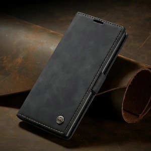 Puede incluir: Una funda de teléfono rectangular de color gris carbón con acabado mate y costuras visibles en los bordes. La funda presenta un pequeño emblema metálico circular con un diseño de estrella situado cerca de la esquina inferior de la tapa.