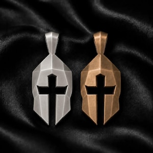 May include: Two metal pendants shaped like Spartan helmets, one in a silver tone and one in a bronze tone. Each helmet features a geometric, faceted design with a central cross-shaped cutout where the eye slit would be, and a top bail for hanging.