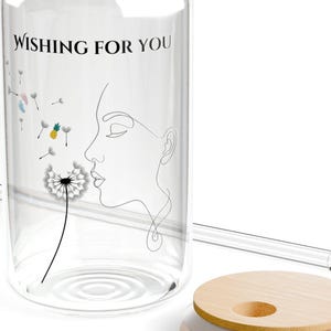 Könnte beinhalten: Ein durchsichtiges, dosenformiges Glas mit Bambusdeckel und Glasstrohhalm. Das Glas zeigt eine schwarze Strichzeichnung eines Gesichtsprofils, das auf eine Pusteblume bläst, sowie den Text "WISHING FOR YOU" und kleine farbige Akzente.