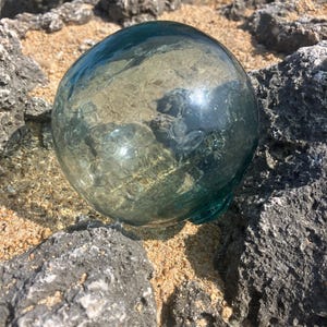 Puede incluir: Una boya de pesca esférica de vidrio con un tono azul verdoso translúcido y una superficie texturizada que presenta numerosas pequeñas burbujas de aire circulares atrapadas dentro del vidrio, creando un aspecto moteado distintivo.